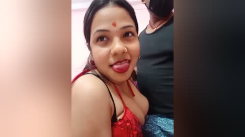 Media: Video of a South Asian woman with medium skin tone, long black hair, and red lipstick, wearing a red lace bra, in a casual setting with a man in a black shirt in the background.