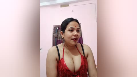 Media: Video of a young, dark-haired South Asian woman with fair skin, wearing a red, sequined halter top, standing in a narrow hallway with pale pink walls and a framed picture.