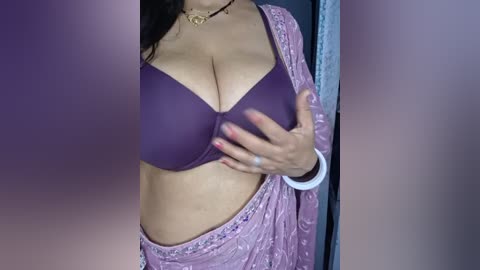 Media: Video of a woman in a purple bra, holding her breasts, with a pink, embroidered sari partially covering her lower body.