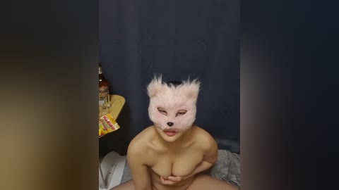 Media: Video of a nude, light-skinned woman with large breasts, wearing a white fox mask, sitting on a bed. Background features a dark curtain and a nightstand with a bottle of alcohol.