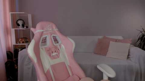 Media: Video of a pastel-pink gaming chair with a cartoon face design, positioned in a minimalist, light grey room with a white sofa and shelves filled with books and decor.