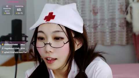 Media: A video of an East Asian woman in a white nurse's cap, glasses, and uniform, speaking into a microphone, with a blurred background of a room.