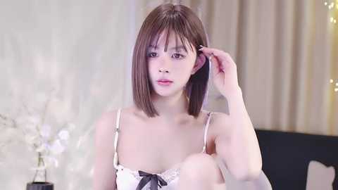 Media: Video of an East Asian woman with straight, shoulder-length brown hair, wearing a white lace bra, adjusting her hair. Background features a white floral patterned wall, a black chair, and a vase of white flowers.