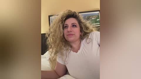 Media: Video of a curly-haired woman with fair skin, wearing a white shirt, lying on a bed with a beige headboard and a framed landscape painting in the background.