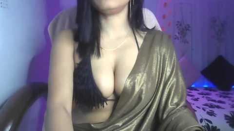 Media: Video of a young woman with long black hair, fair skin, and a low-cut, shimmering gold dress, revealing ample cleavage. Background includes a bed with patterned sheets, and a wall adorned with floral wallpaper.