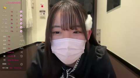 Media: A video of an Asian woman with long black hair, wearing a white surgical mask, black jacket, and a silver chain necklace, indoors with a calendar and white wall background.