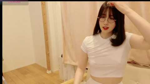 Media: Video of a slender East Asian woman with long black hair, fair skin, and red lipstick, wearing a tight white crop top and white shorts, adjusting her glasses in a beige-tiled bathroom with a wooden floor.