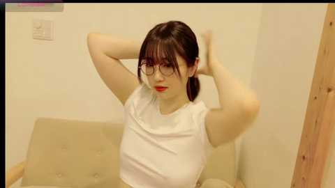 Media: A video of a young Asian woman with pale skin, black hair in a ponytail, wearing glasses and a white t-shirt, adjusting her hair in a beige room.