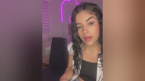 Media: Video of a young Latina woman with medium skin tone and long, wavy black hair, wearing a black top and black-and-white plaid jacket. She is partially hidden by a beige wall. The background features a purple neon light and a white bed with a floral-patterned blanket.