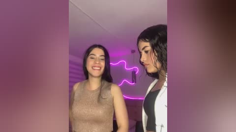 Media: Video of two women with long hair, one in a beige top and the other in a black and white shirt, smiling against a purple-lit wall.