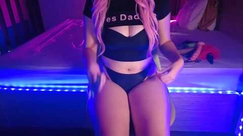 Media: Video of a woman with long pink hair, wearing a black crop top and high-cut black panties, posing seductively in a dimly lit bedroom with purple LED lights.