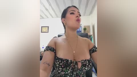 Media: Video of a curvy, light-skinned woman with dark hair, wearing a black floral dress, pouting in a cluttered room with framed certificates and a desk.