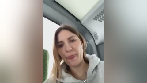 Media: Video of a young Caucasian woman with light brown hair, wearing a grey hoodie, making a funny face while sitting in a car.