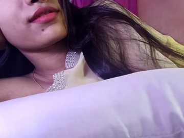 Media: Video of a woman with fair skin, long dark hair, and a mole on her chin, lying on a white pillow, wearing a white top with a sparkly necklace. Pink background.