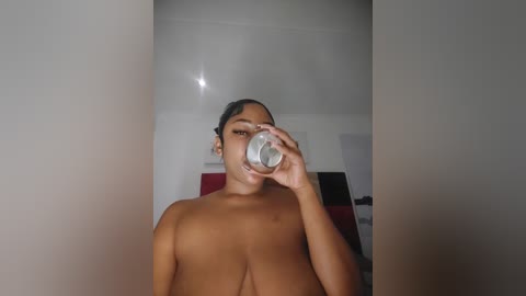 Media: Video of a topless, light-skinned woman with medium-length dark hair, drinking from a clear glass. Background features a white wall and a red and black patterned object.