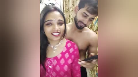 Media: Video of a smiling South Asian woman in a pink saree, holding a phone, with a shirtless man behind her, both indoors.