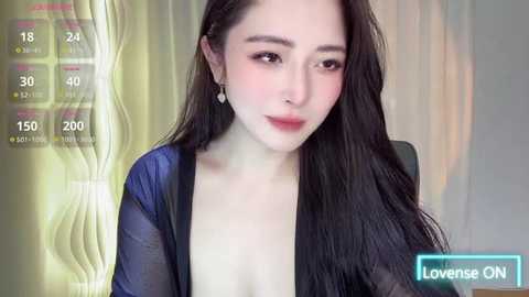 Media: Video of an East Asian woman with long black hair, fair skin, and a slender physique, wearing a sheer blue top, sitting indoors with a yellow and white patterned wall behind her.