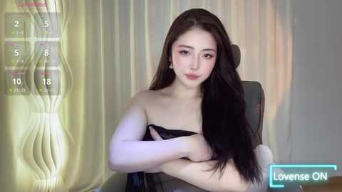 Media: A video of an East Asian woman with long black hair, wearing a black strapless top, sitting on a gray chair. Background shows a yellow curtain. The image includes a live stream overlay.