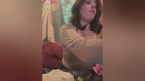 Media: A video shows a woman with shoulder-length brown hair, wearing a beige sweater and floral skirt, sitting on a red sofa, playing with a pink toy.