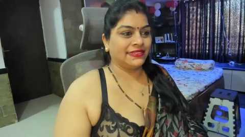 Media: Video of a South Asian woman with medium skin tone, dark hair, and traditional red bindi, wearing a black lace bra and floral sari, seated in a modern bedroom with a bed and window curtains.