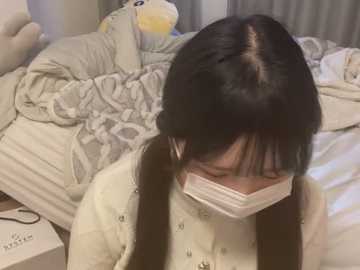 Media: A video of a young Asian woman with long black hair, wearing a white face mask, sitting on a bed covered with a gray and white quilt. The room has a soft, cozy ambiance.