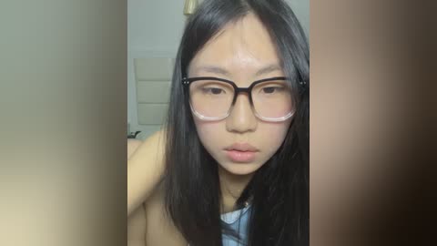 Media: A video of a young Asian woman with long black hair, wearing glasses, and a light blue top, looking introspective in a dimly lit room.