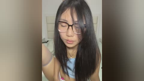 Media: Video of an Asian woman with straight black hair, wearing glasses and a blue crop top, looking downward in a bedroom with beige walls and a white bed.