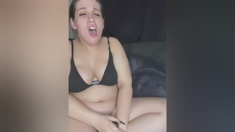 Media: Video of a plus-sized, light-skinned woman with dark hair, wearing a black bikini, sitting on a black couch, mouth open in an expression of pleasure.