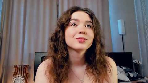 Media: Video of a young woman with curly brown hair and light skin, smiling, indoors with beige curtains and a white lamp in the background.