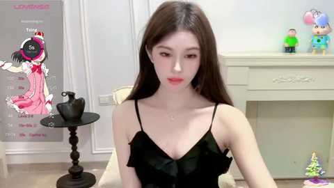 Media: Video of a young Asian woman with long brown hair, wearing a black sleeveless dress, seated indoors with a calendar, toys, and a decorative stand in the background.