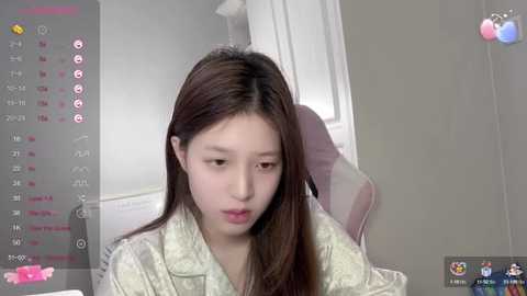 Media: A video of a young East Asian woman with long brown hair, wearing a light green floral-patterned shirt, sitting on a pink chair in a modern room.