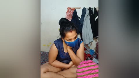 Media: Video of a South Asian woman with medium brown skin and dark hair in a bun, wearing a blue mask and sleeveless top, sitting on a colorful rug in a cluttered room with hanging clothes and a pink striped blanket.