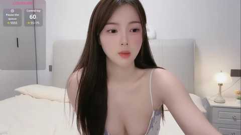 Media: Video of an East Asian woman with long black hair, fair skin, wearing a low-cut white camisole, sitting on a bed in a modern, minimalist bedroom with white walls, a nightstand, and a lamp.