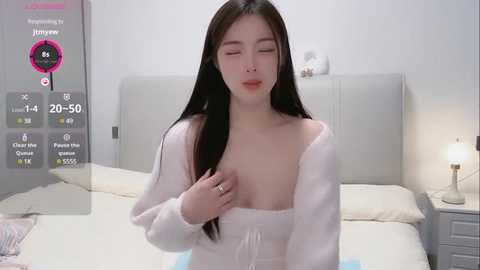 Media: Video of an East Asian woman with long black hair, fair skin, and small breasts, wearing a white robe, in a modern bedroom with a white headboard and nightstand lamp.