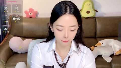 Media: Video of an East Asian woman with straight black hair, wearing a white shirt, sitting on a brown leather couch with stuffed toys, including a yellow duck, a pink star, and a white bear.