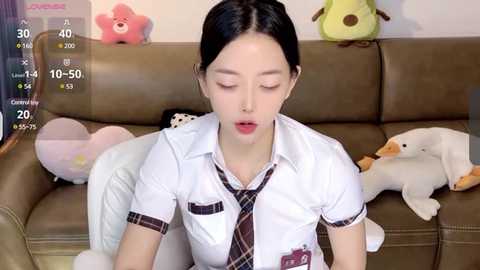 Media: Video of an East Asian woman in a white schoolgirl outfit with a plaid tie, sitting on a brown leather couch. Background features stuffed animals and a screen displaying game statistics.