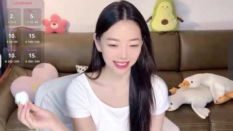 Media: Video of an East Asian woman with long black hair and fair skin, wearing a white top, sitting on a brown leather sofa. Background includes plush toys, including a green frog and a white cat.