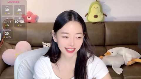 Media: A video of an East Asian woman with long black hair and light skin, wearing a white T-shirt, smiling on a brown couch with plush toys, including a yellow duck, pink bear, and a white and orange penguin.