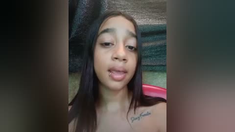 Media: Video of a young, light-skinned Black woman with long, straight black hair, wearing a sleeveless top. She has a tattoo on her left shoulder and is licking her lips. Background includes a green couch and a black blanket.