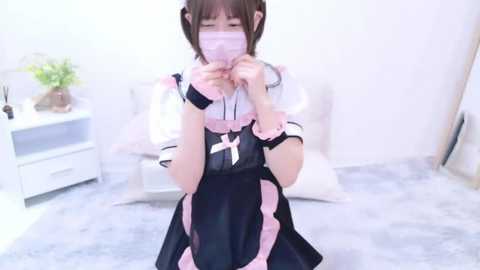 Media: Video of a young Asian woman in a frilly black and pink maid outfit with a pink face mask, kneeling on a light grey carpet in a minimalist room with white walls and furniture.