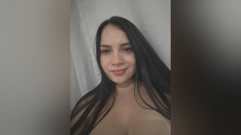 Media: A video of a young woman with long black hair and light skin, smiling softly, standing in front of a plain white background. She appears topless.