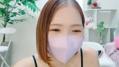 Media: Video of an East Asian woman with straight, shoulder-length brown hair, wearing a pink face mask, black spaghetti strap top, and a gold necklace, in a modern, bright room with white walls, green plants, and white furniture.