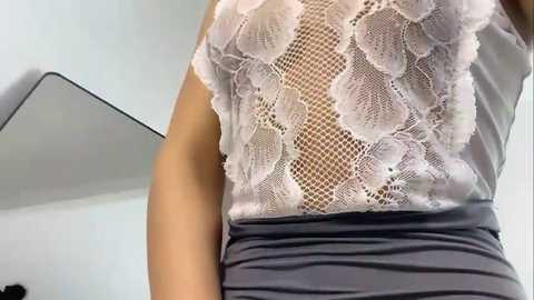 Media: Video of a woman in a sleeveless, white lace top with floral patterns, over a gray, ruched skirt, standing indoors with a mirror and white wall in the background.