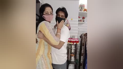 Media: Video of a young South Asian woman in a yellow and pink dress, with a man in a white shirt, both wearing masks, hugging in a kitchen with stainless steel appliances and colorful utensils.