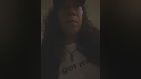 Media: Video of a woman with curly hair and a black cap, wearing a white t-shirt with \"got milk?\" text, and a necklace, standing against a dimly lit, dark background.