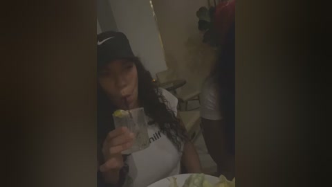 Media: A dimly lit video of a young woman with long hair, wearing a black cap and white t-shirt, drinking a clear liquid from a plastic cup. Background includes a bed and blurred objects.