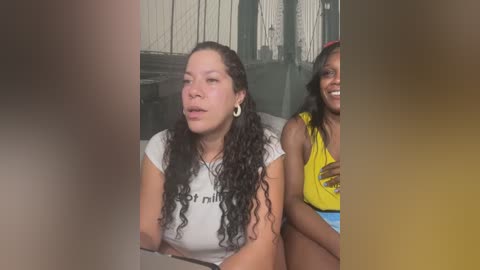 Media: Video of two women in casual attire, one with long curly hair, the other with straight hair, sitting side by side in a dimly lit room with white wooden paneling.