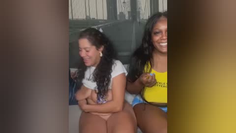 Media: Video of two women with medium to dark skin tones. One with long curly hair, white crop top, and pink panties; the other with straight hair, yellow crop top, and blue shorts. Both smiling. Background shows a modern room with a glass door.