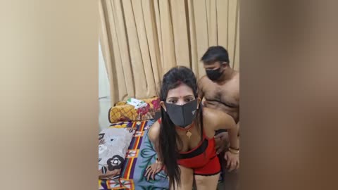 Media: A video shows a topless South Asian man with a mask, standing behind a woman with a mask, in a room with beige curtains and colorful bedding.