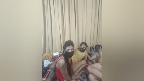 Media: A video shows three individuals, a woman with long hair, a man with glasses, and another man, all wearing masks, sitting on a bed with a yellow blanket in a beige room.